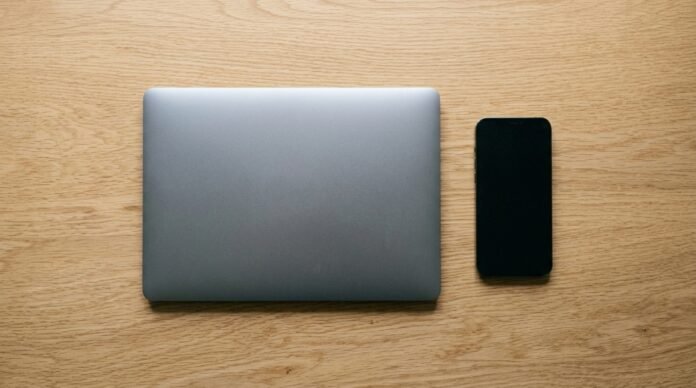 A smartphone and a laptop side by side on a desk, each showing a blank screen, soft daylight.