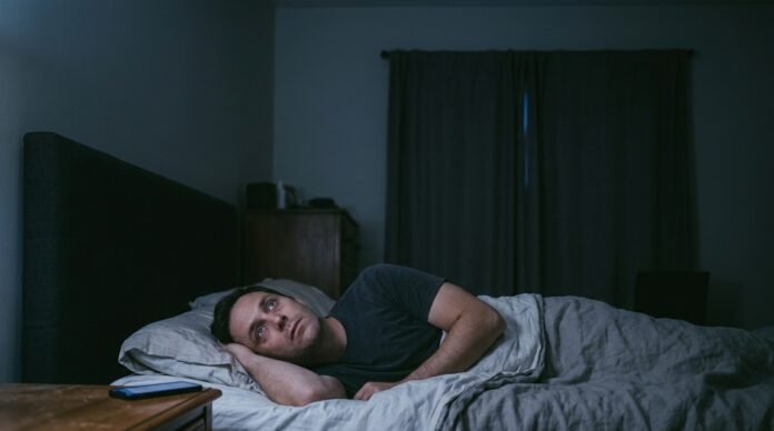 A person lying awake in bed at night, eyes open, faint light from a phone or window across the dark room.