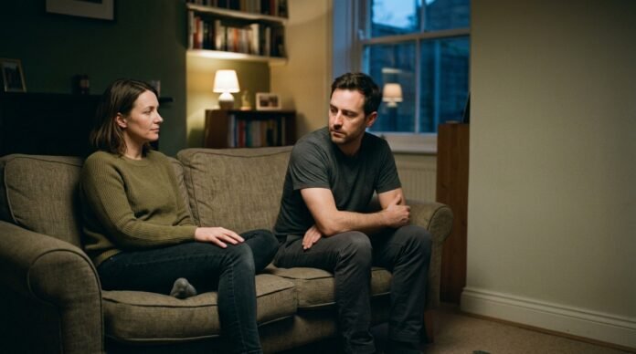 Two people sitting on a sofa at a slight distance, one turned toward the other who is looking away, in calm evening light.