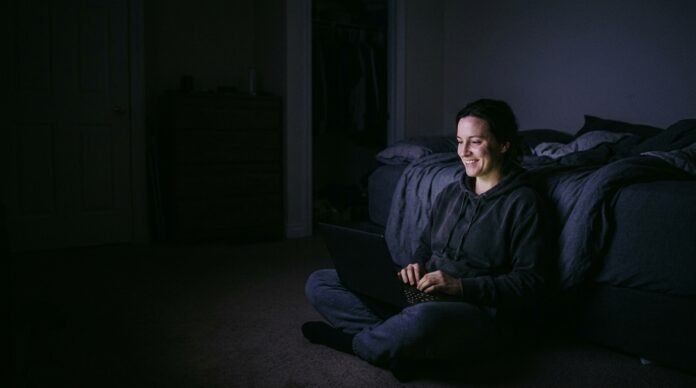 A person sitting on the floor of a bedroom late at night, laptop open, mid-video call, lit only by the screen.