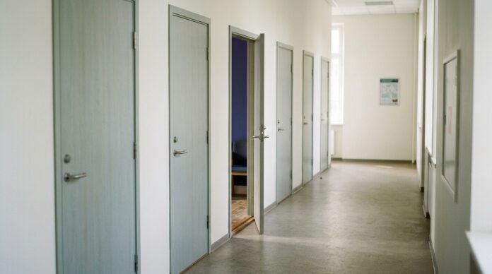 Several similar-looking closed doors along a plain corridor in soft daylight, one slightly ajar.