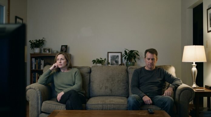 Two people sitting on the same sofa in the evening, both looking at the room rather than each other, in quiet lamplight.