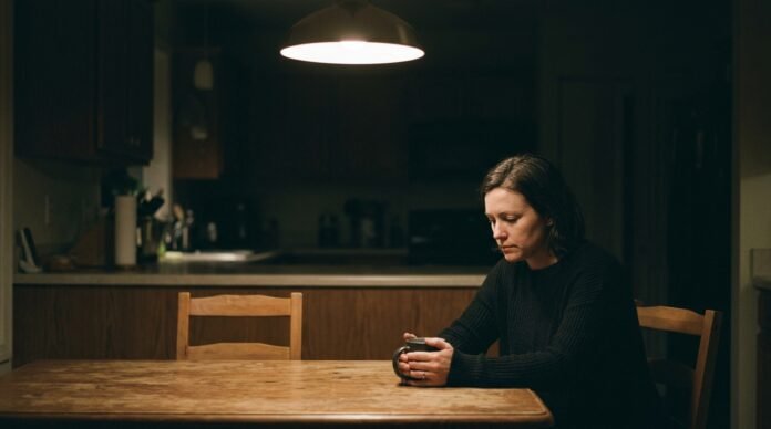 A person sitting alone at a kitchen table at night, hands around a mug, looking thoughtful and unsettled.