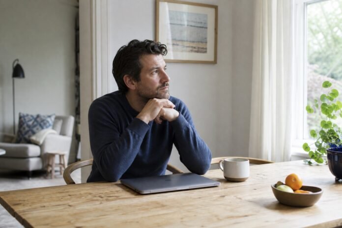 A man sits in daylight at a dining table beside a closed laptop, looking across an unstructured day.